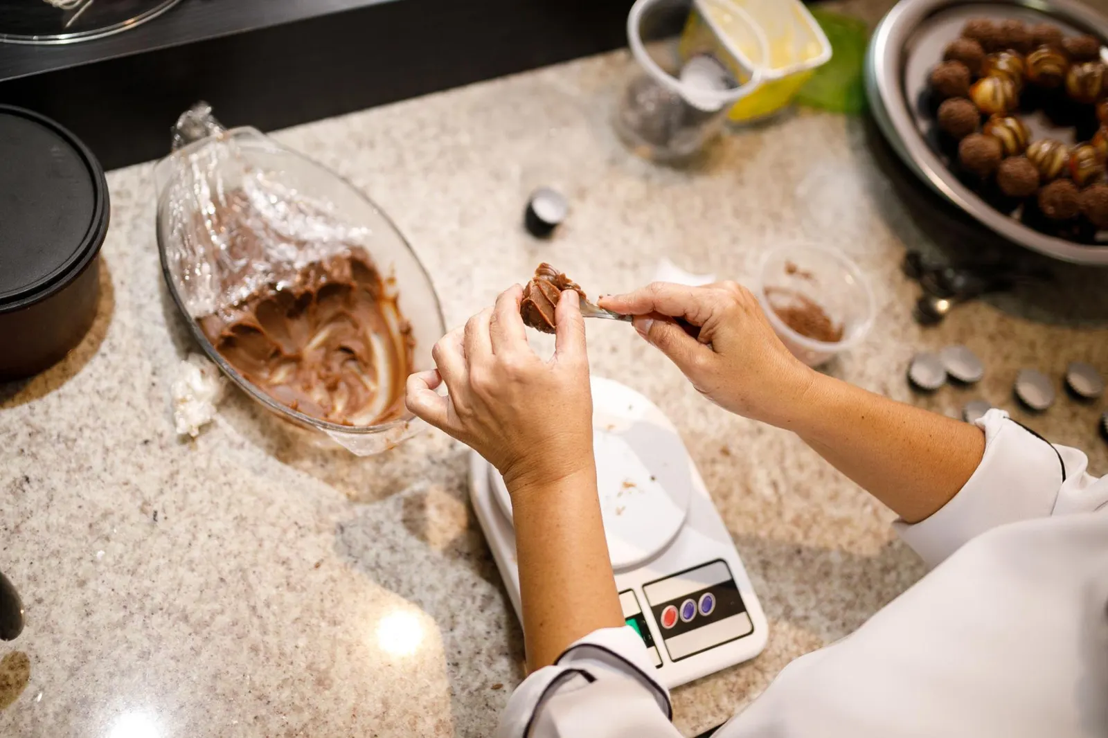 Mãos modelando um brigadeiro sobre balança digital em uma bancada de confeitaria